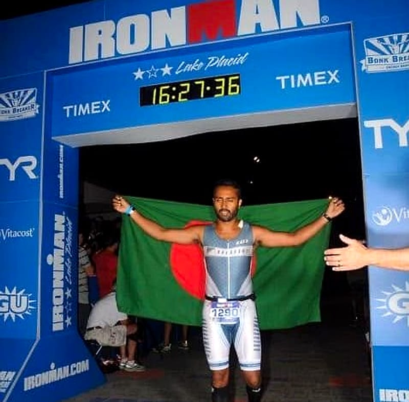 Tahsin Alam holds up the flag of Bangladesh as he crosses the IRONMAN Lake Placid finish line, the clock reading 16:27:36